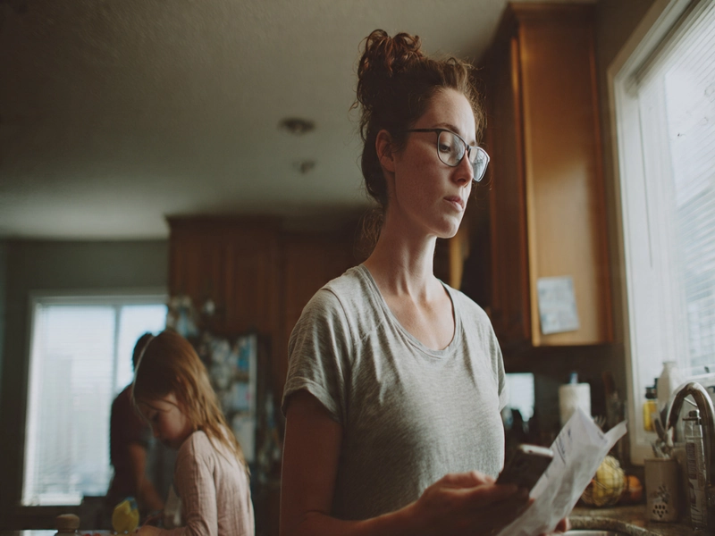Parent standing in the kitchen reading paperwork while mentally overwhelmed, with a child in the background needing attention